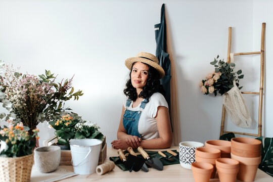 Young female horticulturist in straw hat sitting near flowers on table with assorted tools at home