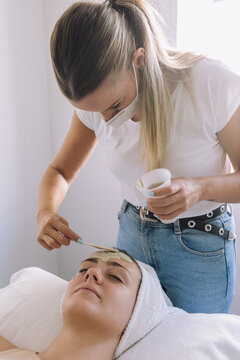 Cosmetician applying alginate mask on face of anonymous young female customer lying on couch with closed eyes