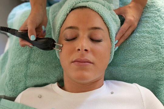 Top view of crop unrecognizable female cosmetologist doing rejuvenate facial procedure with microcurrent device for graceful lady lying on couch with closed eyes