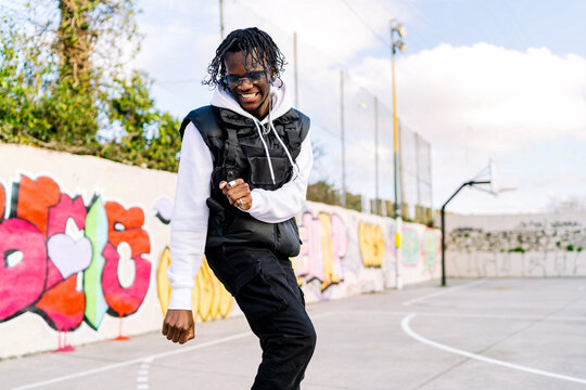 Young trendy ethnic man in sunglasses dancing on tiptoes while looking down against graffiti wall in city