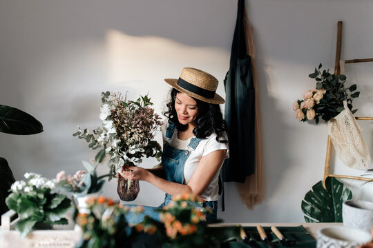 Young female horticulturist in straw hat creating bouquet on table with assorted tools at home