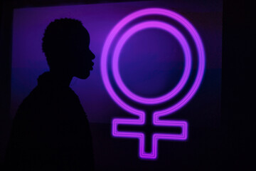 Side view of unrecognizable trendy black model silhouette standing near neon female symbol glowing with purple light