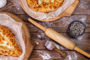 Khachapuri lies on wooden round pizza boards, chicken eggs and a rolling pin on a brown board background.