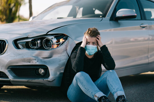 Sad young female driver in casual clothes and medical mask sitting on road near expensive damaged car and crying after accident during coronavirus outbreak