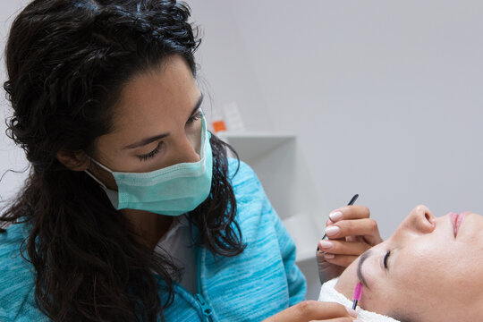 Crop anonymous beautician in mask combing eyebrow of adult woman with closed eyes using brush during facial procedure in beauty center