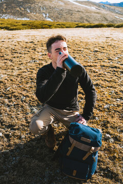 From above young stylish male traveler in casual outfit sitting on haunches near backpack and drinking hot beverage from thermos during hiking trip in mountainous terrain