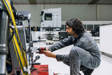 Side view of male mechanic in dirty uniform using electric screwdriver and fixing wheel of custom motorbike in bright workshop