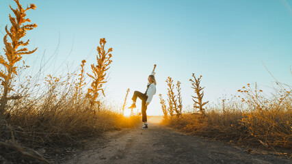 Side view of active young lady in trendy outfit dancing in dry fiends against cloudless blue sky in countryside