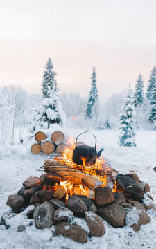 Teapot placed on bonfire on snowy field near coniferous trees against cloudy sunset sky in winter