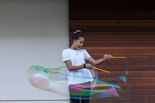 Cheerful young African American female teenager with dark hair in white t shirt playing with big colorful soap bubbles and having fun standing on street near wooden wall