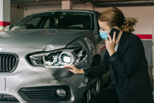 Unrecognizable concentrated female insurance officer in formal suit and protective mask talking on mobile phone while inspecting car damages and filling out claim after accident