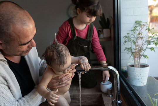Little girl washing cute baby in arms of father during bathing in sink in kitchen