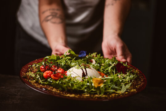 Hands of anonymous person holding a yummy burrata cheese on cold tomato cream with arugula leaves and cherry tomatoes with truffles and peanuts