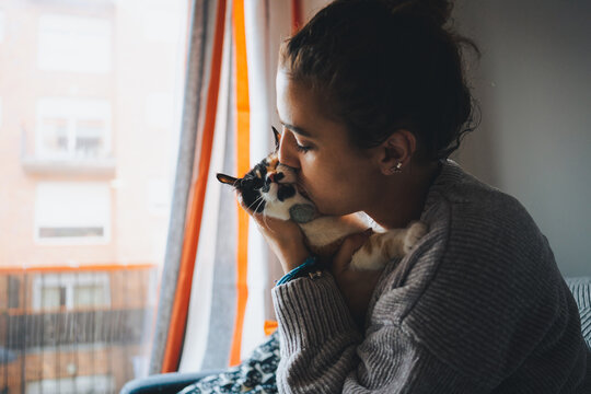 Side view of young female owner in casual outfit hugging and kissing cute fluffy calico cat with closed eyes while resting on sofa in living room