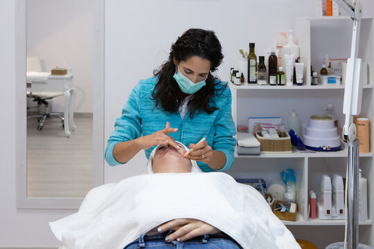 Unrecognizable beauty master applying hot wax with spatula on face of lying woman during epilation process in spa salon
