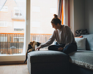 Side view of young lady in casual clothes sitting on comfortable couch with crossed legs and playing with adorable calico cat in modern apartment
