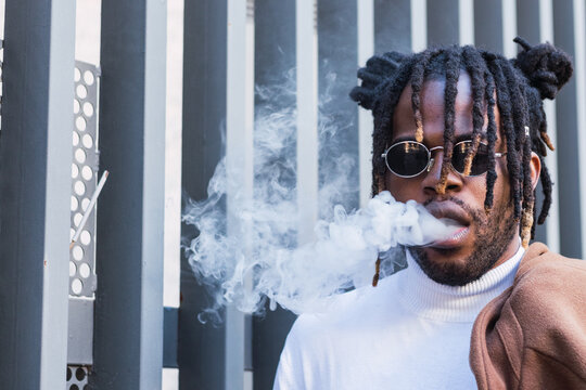 Modern adult hipster African American male in trendy outfit and in sunglasses with dreadlocks smoking electronic cigarette standing on the street