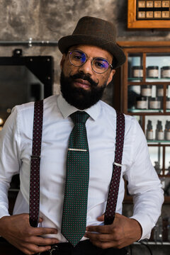 Crop bearded dandy ethnic male hairdresser in eyeglasses with mustache standing looking at camera in barbershop