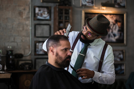 Stylish dandy serious ethnic male barber trimming hair of adult client with electric clipper in hairdressing salon