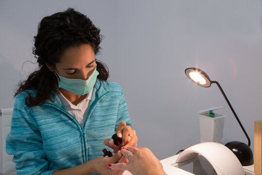 Attentive female manicurist in face mask applying lacquer on nail of crop woman near shiny lamp in beauty salon