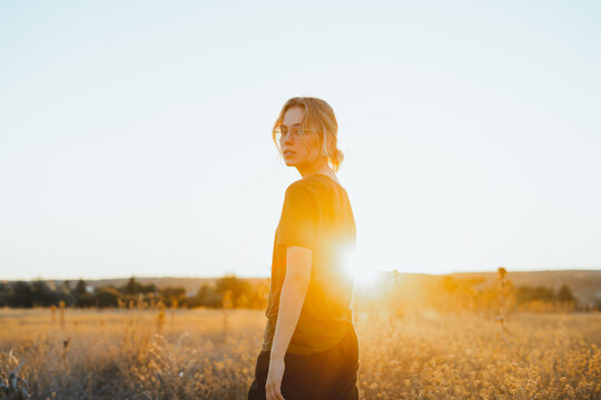 Side view of confident young female traveler in casual clothes and eyeglasses standing in meadow and looking t camera while relaxing in countryside at sunset