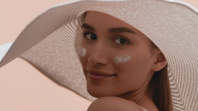 Young good-looking dark-haired European woman in a big white hat with smears of sun cream on her cheeks turns to the camera smiling against beige background | Sunblock commercial