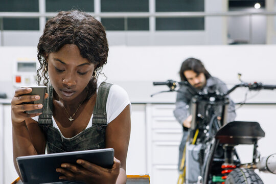 Busy African American female mechanic using tablet and drinking coffee on background of male technician fixing motorcycle in garage