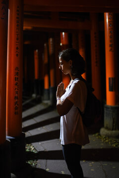 Young caucasian woman prays with hands together at Fushimi Inari Shrine (Fushimi Inari Taisha) surrounded by vermilion torii gates at night, Kyoto, Japan