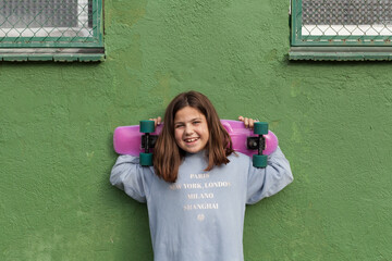 Happy teenage girl in casual outfit and sneakers standing near aged green building with skateboard behind head after riding