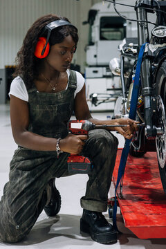 Side view of skilled African American female mechanic in protective headphones using power screwdriver while assembling custom motorcycle in workshop