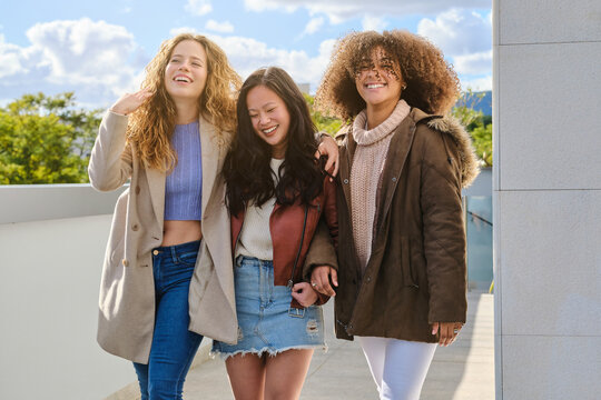 Group of cheerful millennial multiracial girlfriends in casual warm clothes hugging and chatting friendly while enjoying time together on terrace of urban building