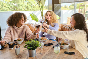Cheerful multiracial young women in casual clothes toasting with glasses of wine while gathering at table and having dinner together in restaurant