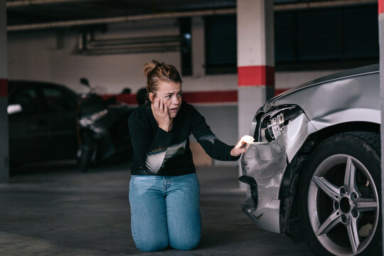Upset young lady in casual clothes sitting on knees and touching damaged car headlights after accident in parking space