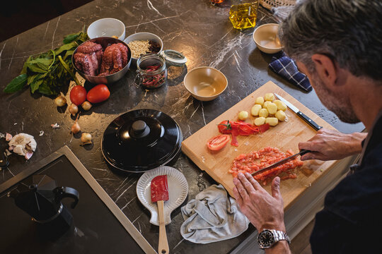 From above anonymous male cutting fresh tomato on board near peeled potatoes on table near bowls with squid and grain and heap of bay leaves and tomatoes with onions and garlic and jar of chili peppers