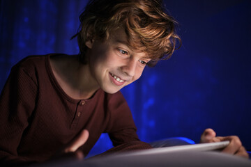 Happy teenage boy lying in bed under blanket and watching video on tablet in dark bedroom with glowing lights