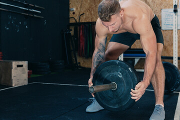 Muscular sportsman with strong naked body putting heavy weight plate on barbell while preparing for functional training in gym
