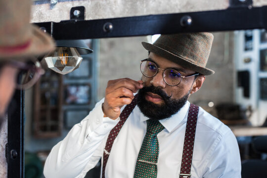 Crop bearded dandy ethnic male hairdresser in eyeglasses reflecting in mirror while curling mustache in barbershop