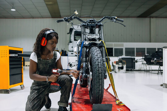 Side view of skilled African American female mechanic in protective headphones using power screwdriver while assembling custom motorcycle in workshop
