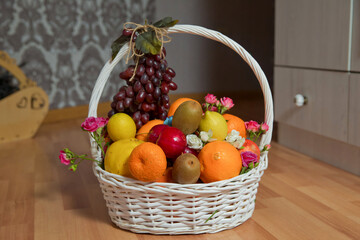 A bunch of grapes hangs from the handle of the basket. The white basket contains grapes, kiwi, orange, red apple, quince, orange, and lemon. There are pink and white golden flowers.
