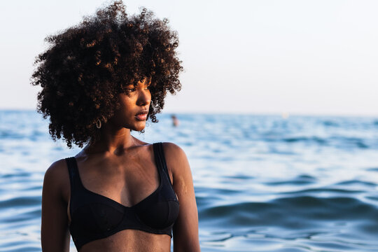 Tranquil black female wearing bikini standing in calm sea water in summer and looking away