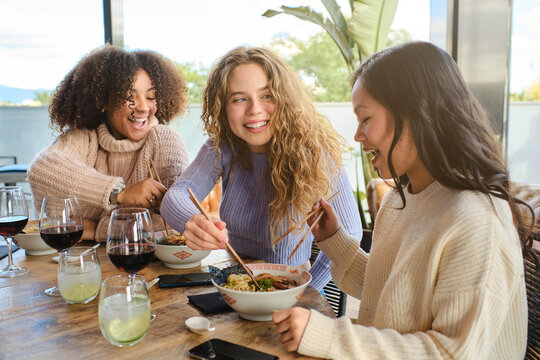 Group of cheerful multiracial young women having fun while eating traditional Asian ramen during dinner in restaurant