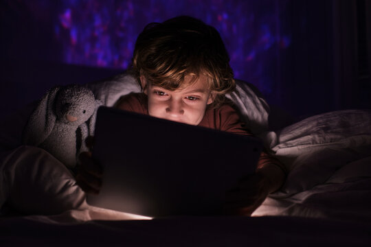 Happy teenage boy lying in bed under blanket and watching video on tablet in dark bedroom with glowing lights