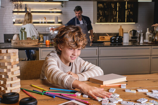 Cheerful boy mixing dominoes near marker pens and book on table against parents cooking lunch in kitchen at home