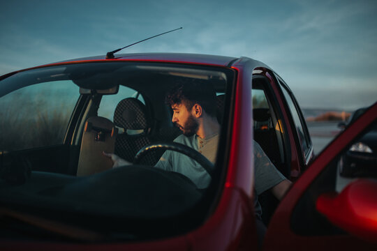 Concentrated young bearded male skater in casual outfit putting skateboard in car parked on asphalt road in nature