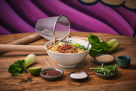 Stock photo of yummy noodles soup with minced meat isolated in wooden background next to sesame seeds and other ingredients.