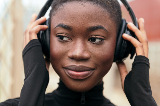 Crop young dreamy black female listening to music from wireless headphones while looking away in daylight