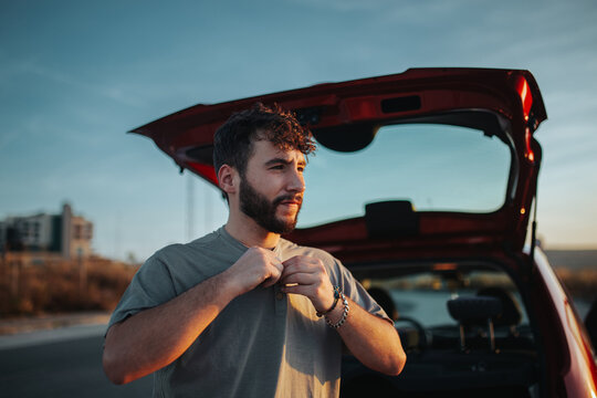 Young bearded male driver sitting in opened car trunk and putting shirt on looking away