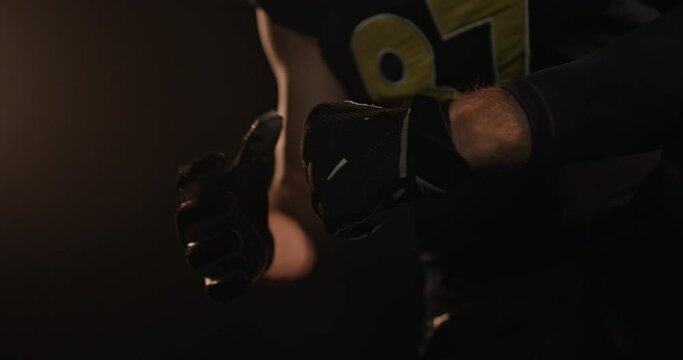 Crop rugby player cracking knuckles in dark studio. Crop unrecognizable male American football player in uniform and gloves sitting in dark studio and cracking knuckles
