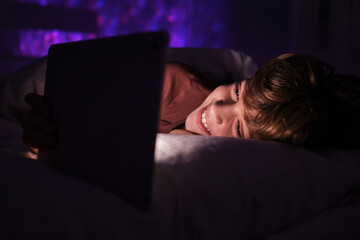 Happy teenage boy lying in bed under blanket and watching video on tablet in dark bedroom with glowing lights