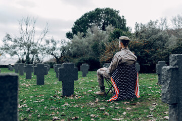 Back view full body of soldier in uniform sitting on chair with American flag while mourning  death of warriors at graveyard